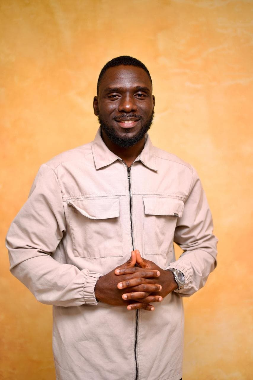 Oyewale Abdullah Muftau smiling, hands clasped, against a warm ochre wall